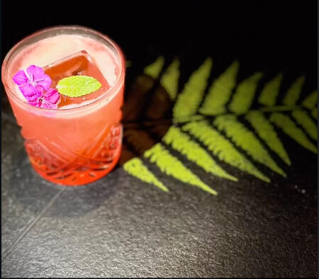 Pink cocktail in a textured glass garnished with a purple flower and mint, placed on a dark surface with a fern leaf casting a shadow beside it.
