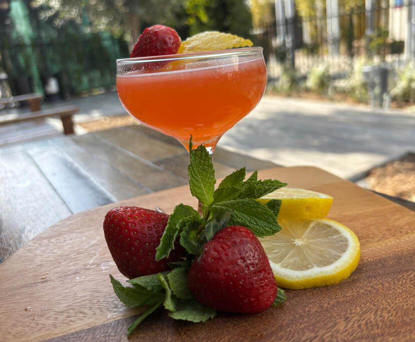 Red-colored cocktail in a coupe glass garnished with strawberries, mint, and lemon, displayed on a wooden surface outdoors.