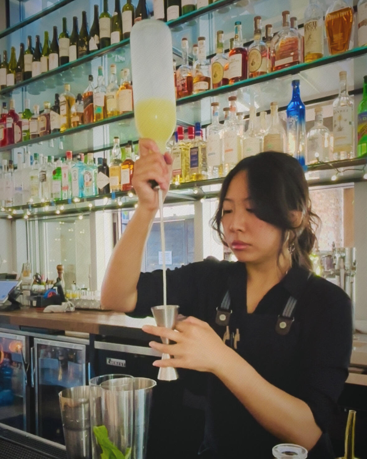 A woman with curtain bangs pouring lemon juice from a long-spout bottle into a jigger behind a bar.